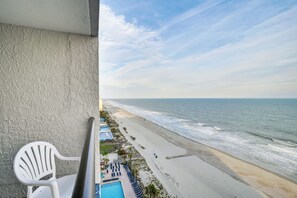Ein Balkon mit weißem Stuhl, Blick auf einen Strand mit Poolbereich und Liegestühlen.