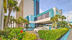 Un hotel moderno con una fachada destacada en azul y blanco, un área de estacionamiento y exuberante jardinería.