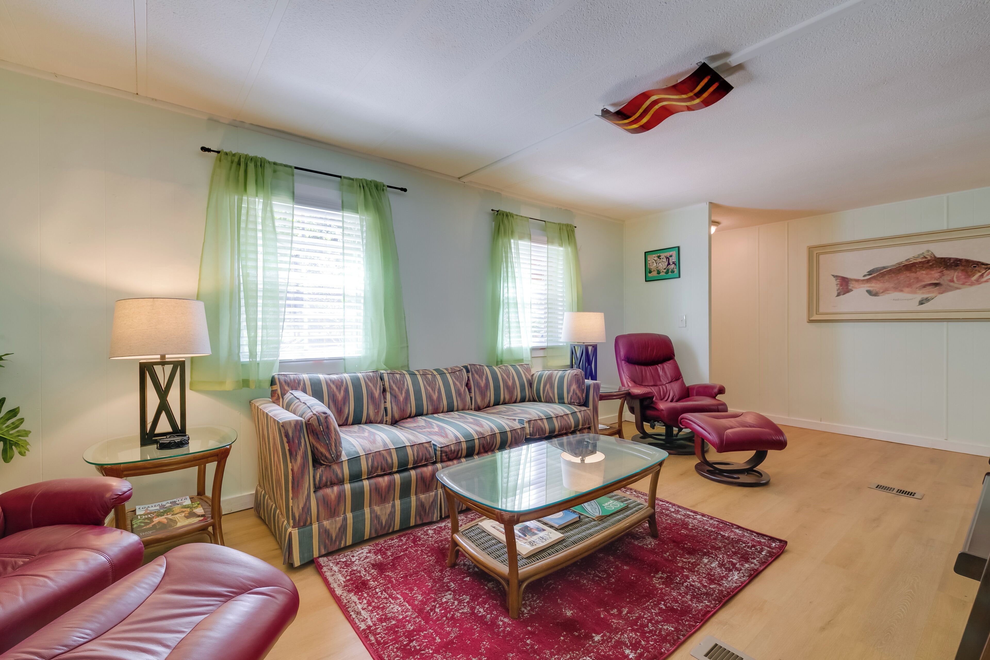 A living room with a striped sofa, a red leather couch, a glass coffee table, and a red rug. There are two lamps, a fish painting, and a ceiling flag.