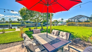 An outdoor patio with a table and chairs, a red umbrella, and a view of a well-maintained lawn and a greenhouse structure in the background. - Punta Gorda Escape w/ Private Lanai & Pool! (Punta Gorda)
