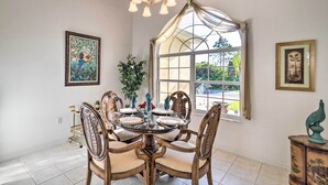 A dining area with a round table, chairs, and a chandelier. There is a window with a view of greenery outside. - Port Charlotte Home on Canal w/ Lanai & Pool! (Port Charlotte)
