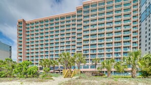 A multi-story hotel with a uniform facade, balconies, and a sandy ground area with palm trees. - Myrtle Beach Oceanfront Condo w/ Pool & Lazy River (Myrtle Beach)