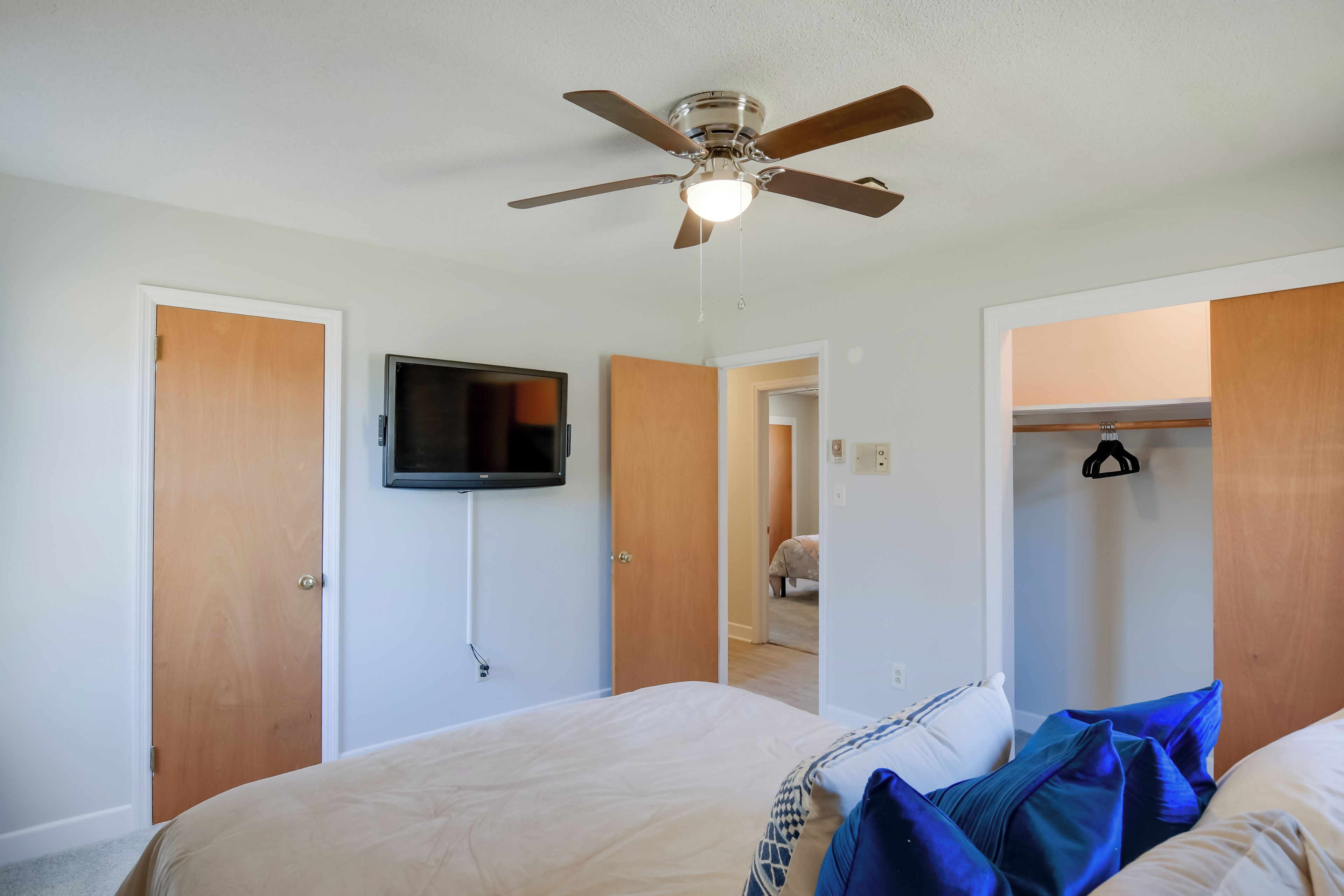 A bedroom with a bed, a ceiling fan, a TV mounted on the wall, and a closet with a hanging rack.