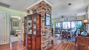A spacious living room with hardwood floors, a brick accent wall, a wooden cabinet, and a dining area with a table and chairs. - Rustic Beaufort Home w/ Stunning Marsh Views! (Beaufort)