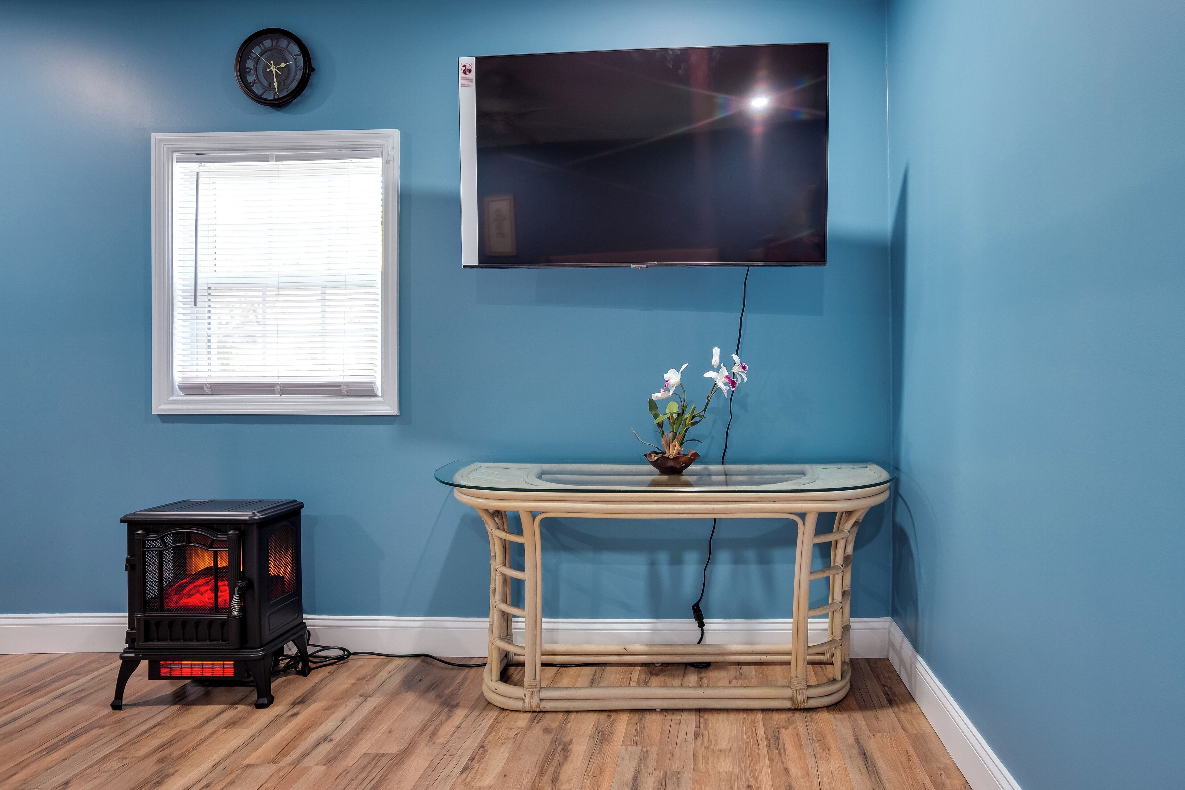 Una habitación con pared azul, un televisor de pantalla plana montado sobre una mesa con cubierta de vidrio, una planta, un calentador pequeño negro y una ventana con persianas.