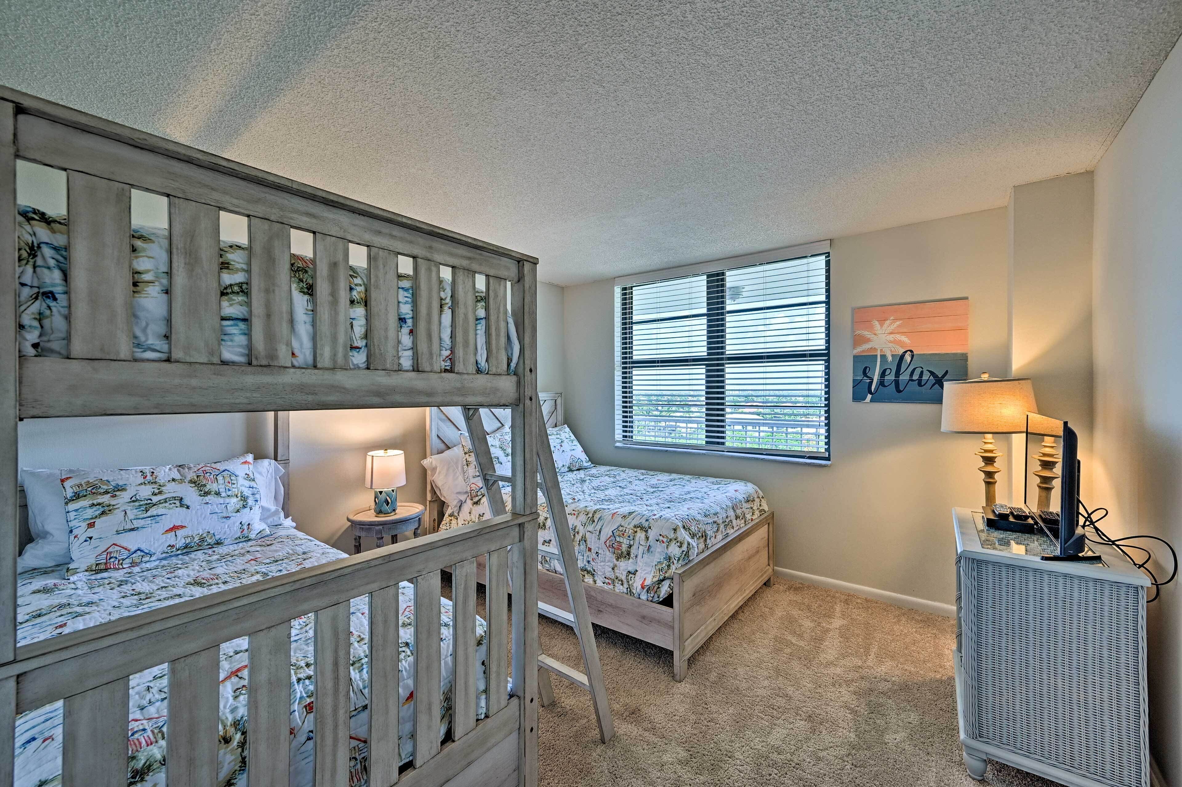 A bedroom with bunk beds, a bedside table with a lamp, a window with blinds, and a wall-mounted TV.