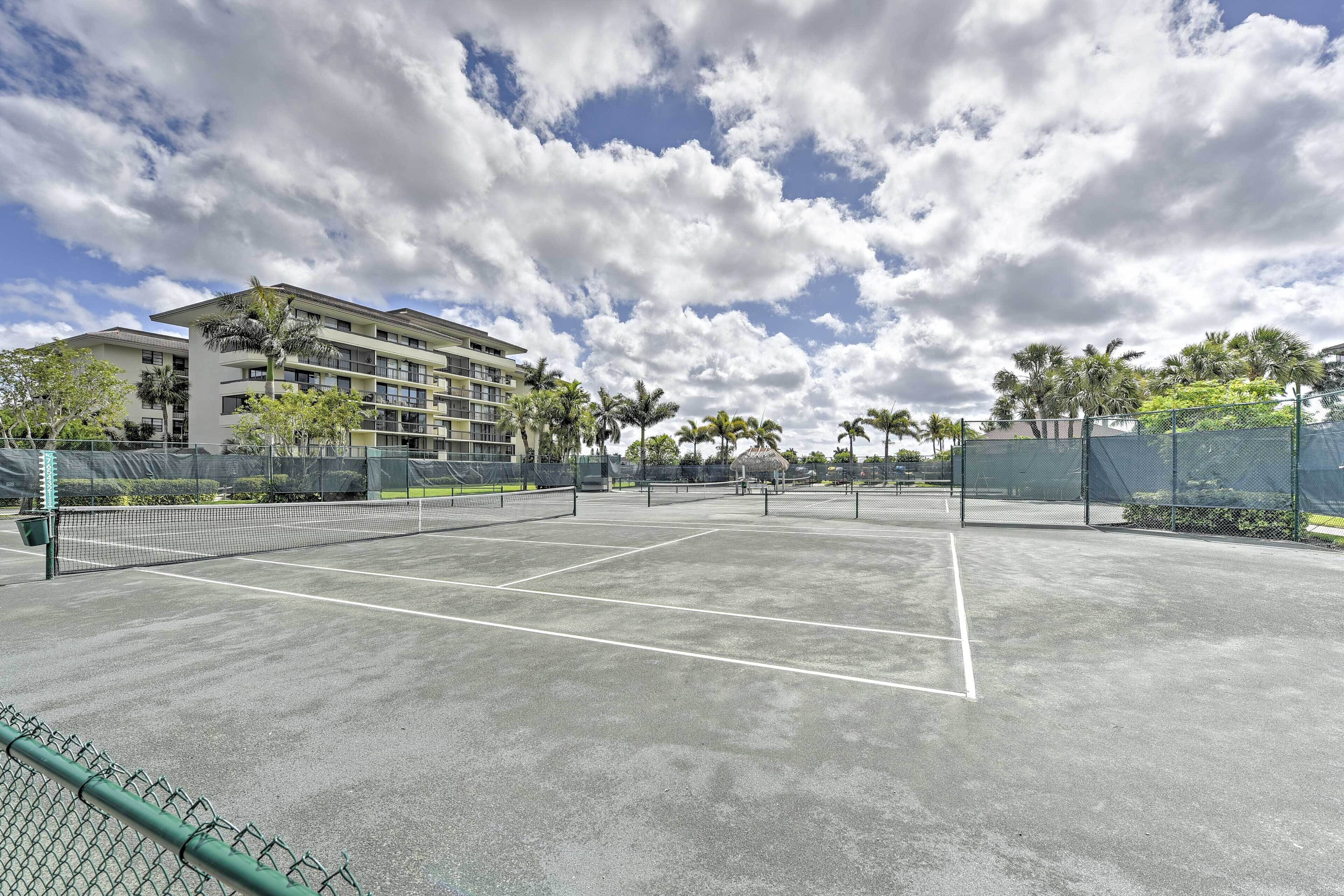Een tennisbaan met net, omringd door een hek, palmbomen en een gebouw op de achtergrond.