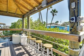 A wooden deck with four stools, a covered area with a roof, and a view of a blue building and palm trees in the background.