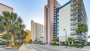 A high-rise hotel with a prominent clock tower, multiple balconies, and a palm tree-lined street. - Oceanfront Resort-style Condo in Myrtle Beach! (Myrtle Beach)