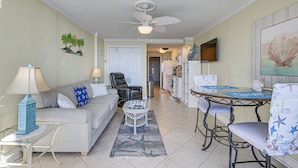 A living room with a sofa, coffee table, and a dining area with a table and chairs. There is a ceiling fan, a TV mounted on the wall, and a kitchen in the background. - Oceanfront Resort-style Condo in Myrtle Beach! (Myrtle Beach)