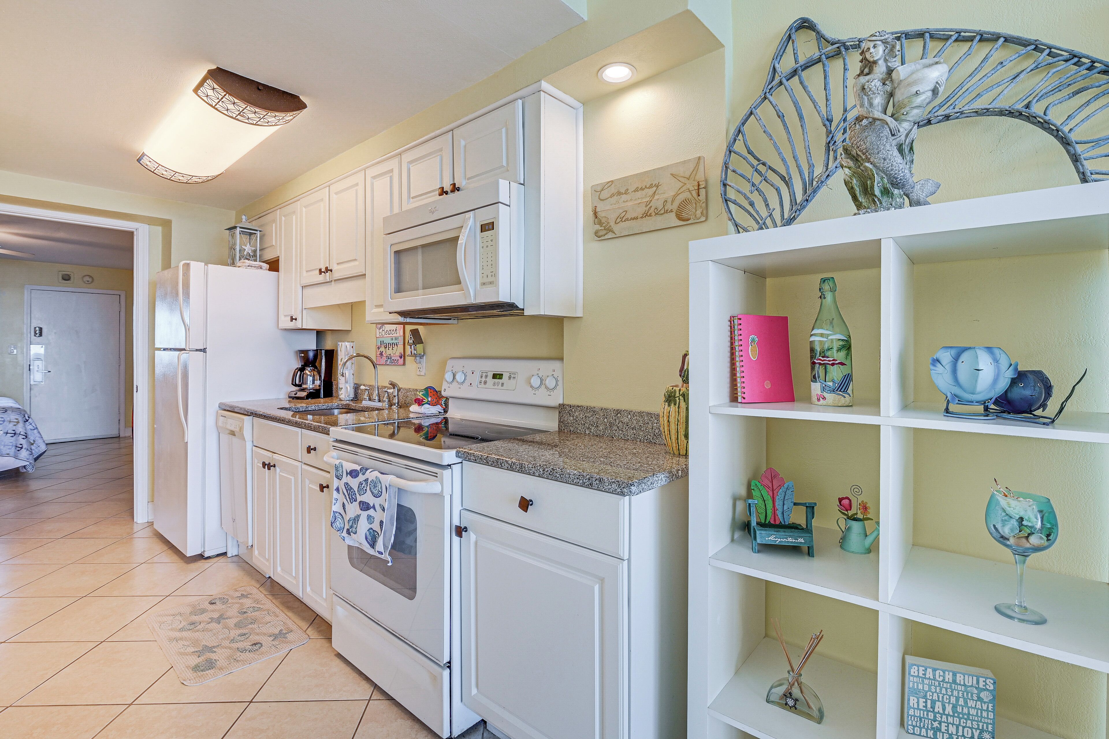 Una cocina con armarios blancos, un microondas y una estufa. Hay objetos decorativos en los estantes y un cartel en la pared.
