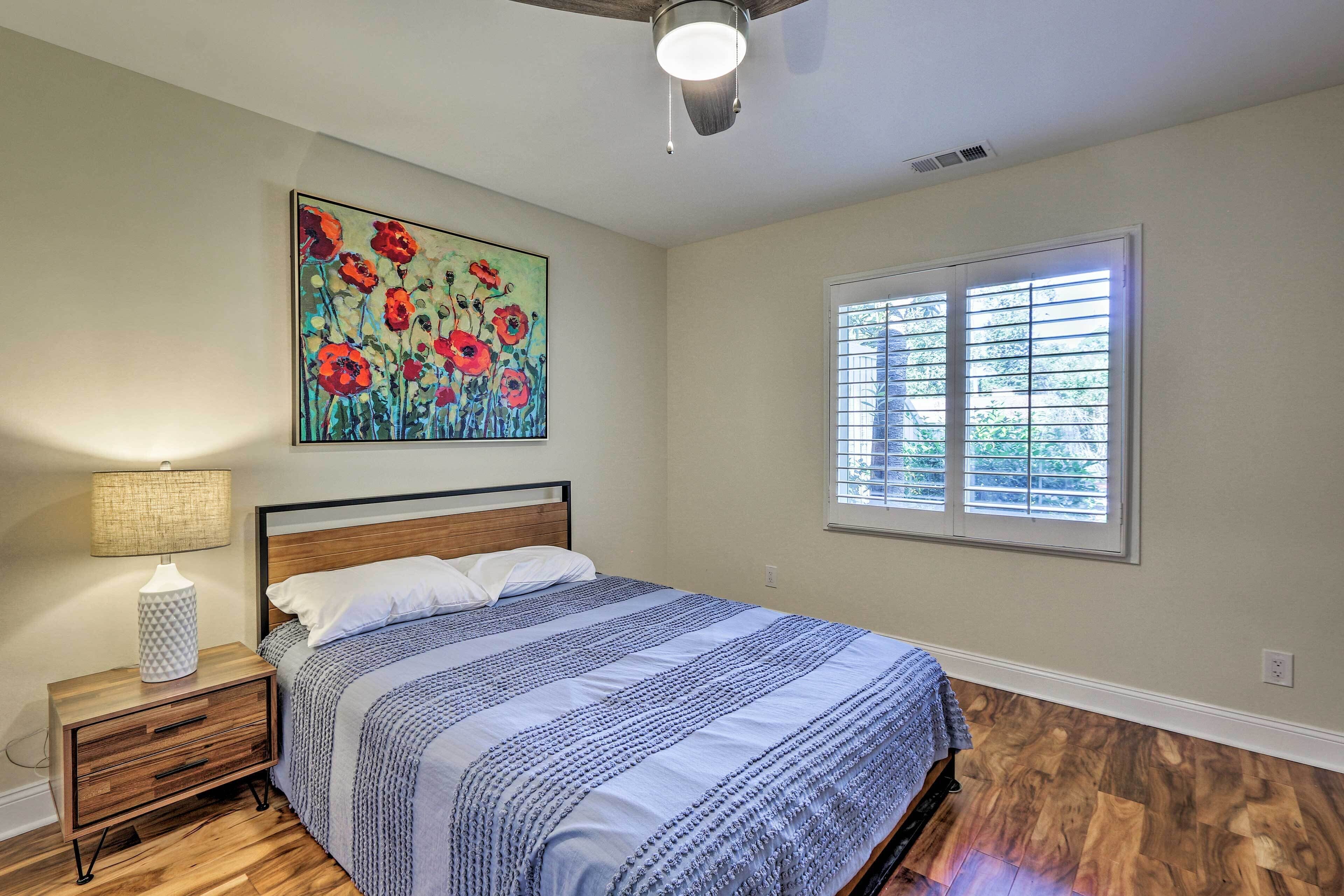 Une chambre avec un plancher en bois, un lit recouvert d’une couvre-lit à rayures bleues et blanches, une table de chevet avec une lampe, un tableau représentant des fleurs rouges et une fenêtre avec des stores.