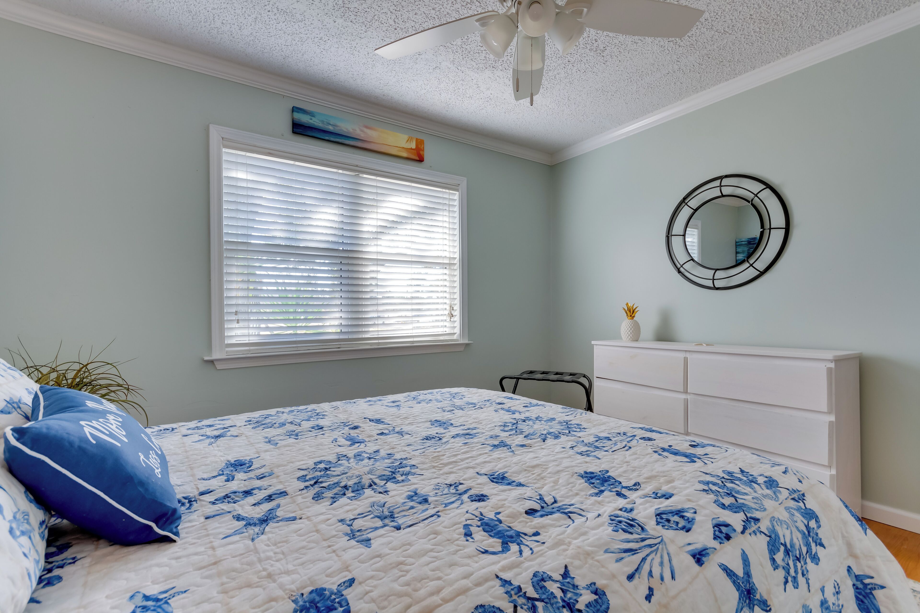 A bedroom with a bed, a ceiling fan, a window with blinds, a dresser, and a mirror on the wall.
