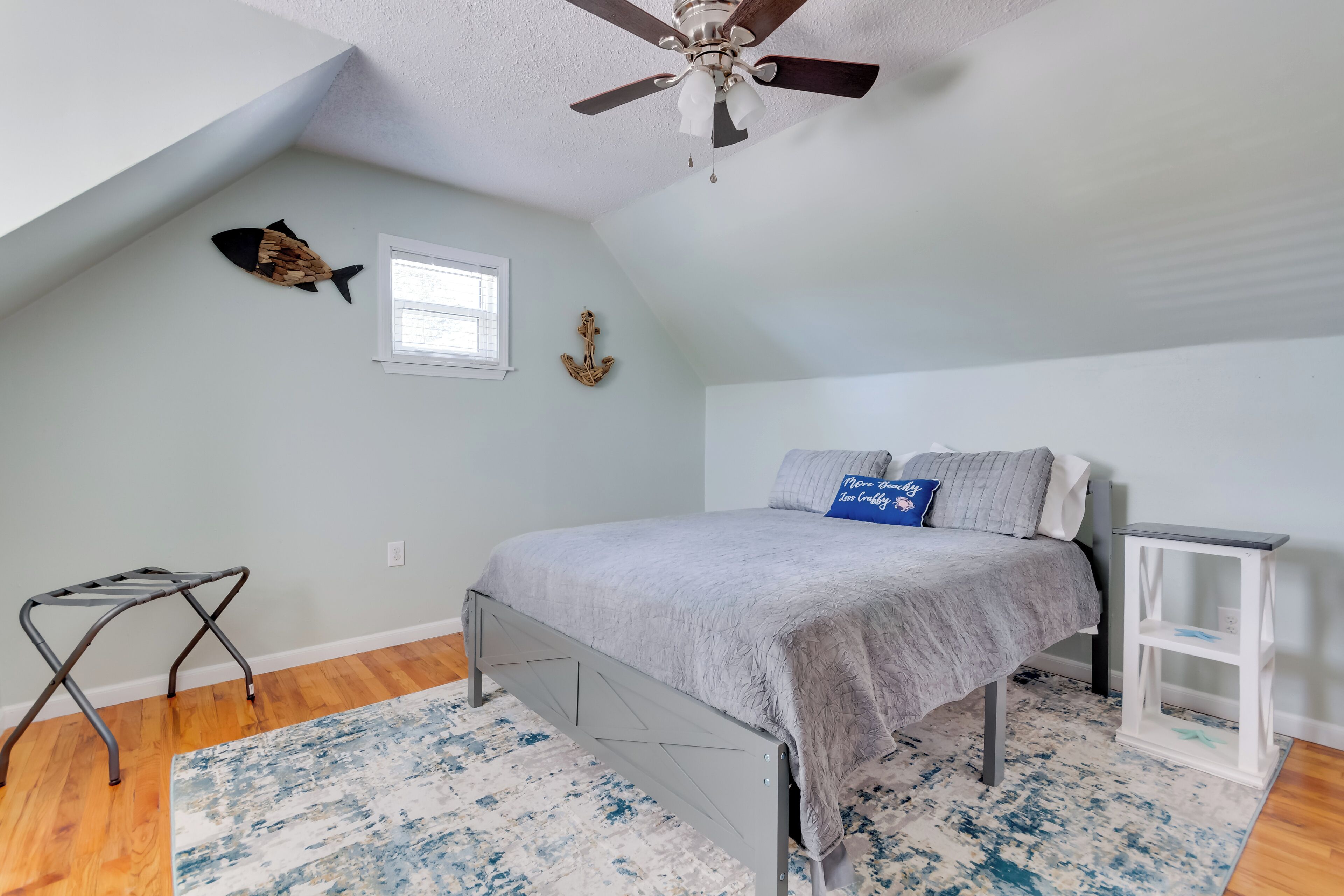 A bedroom with a sloped ceiling, a ceiling fan, a bed with gray bedding, a small window, and a decorative fish on the wall.