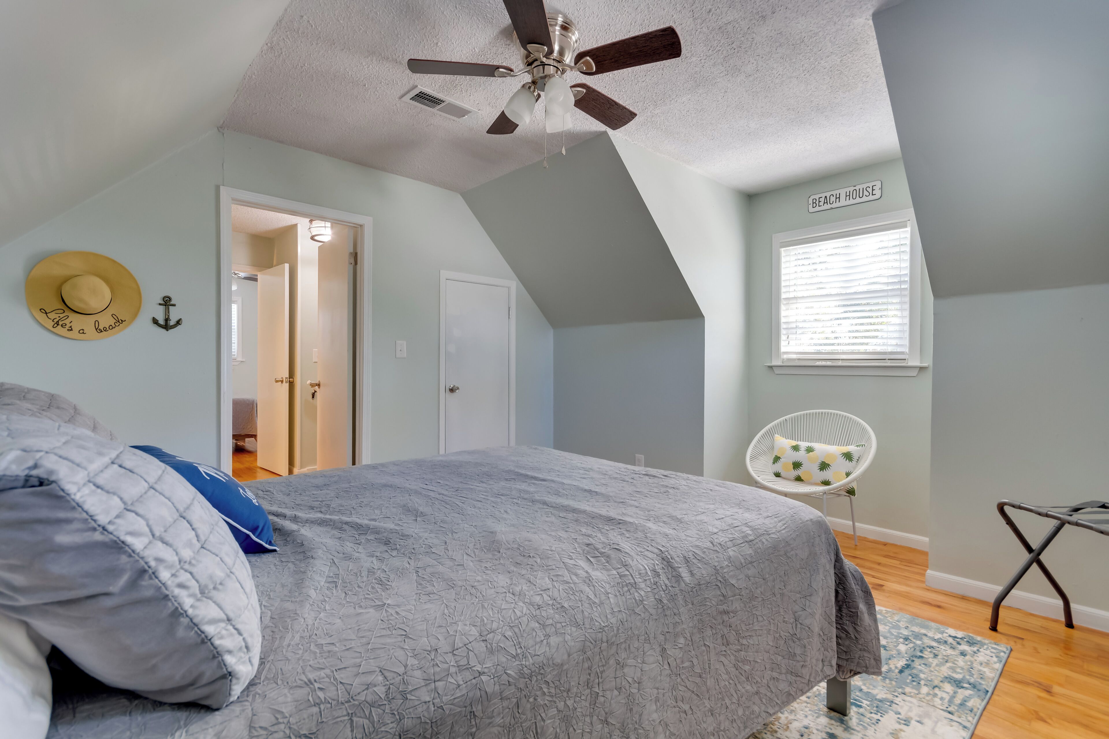 A bedroom with a sloped ceiling, a ceiling fan, a bed with a gray quilt, a window with blinds, and a chair with a cushion.