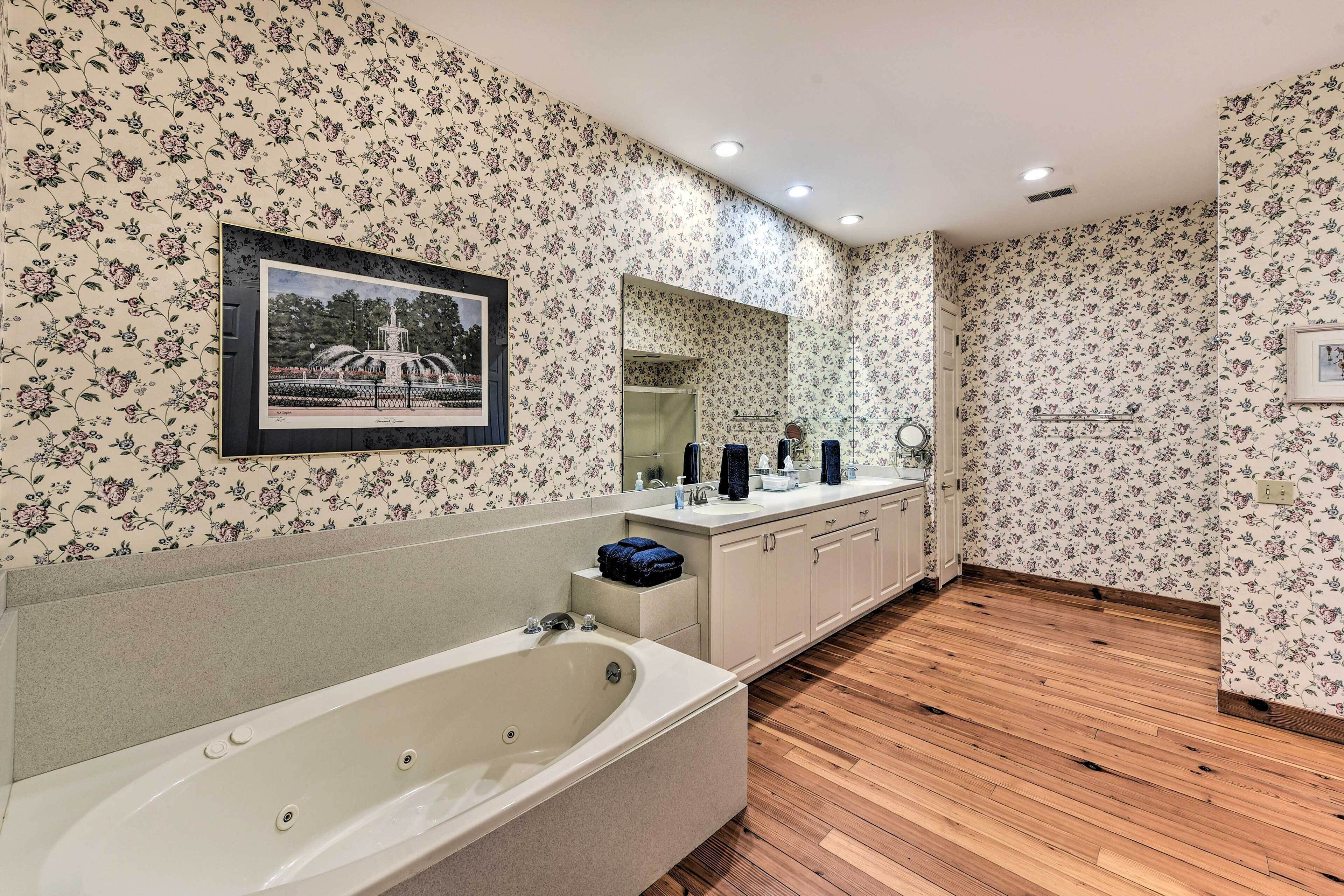 A bathroom with floral wallpaper, a large mirror, a bathtub, wooden flooring, and a framed picture on the wall.