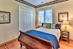 Un dormitorio con cama de madera, ventana con persianas, mesita de noche con lámpara y cuadros en las paredes.