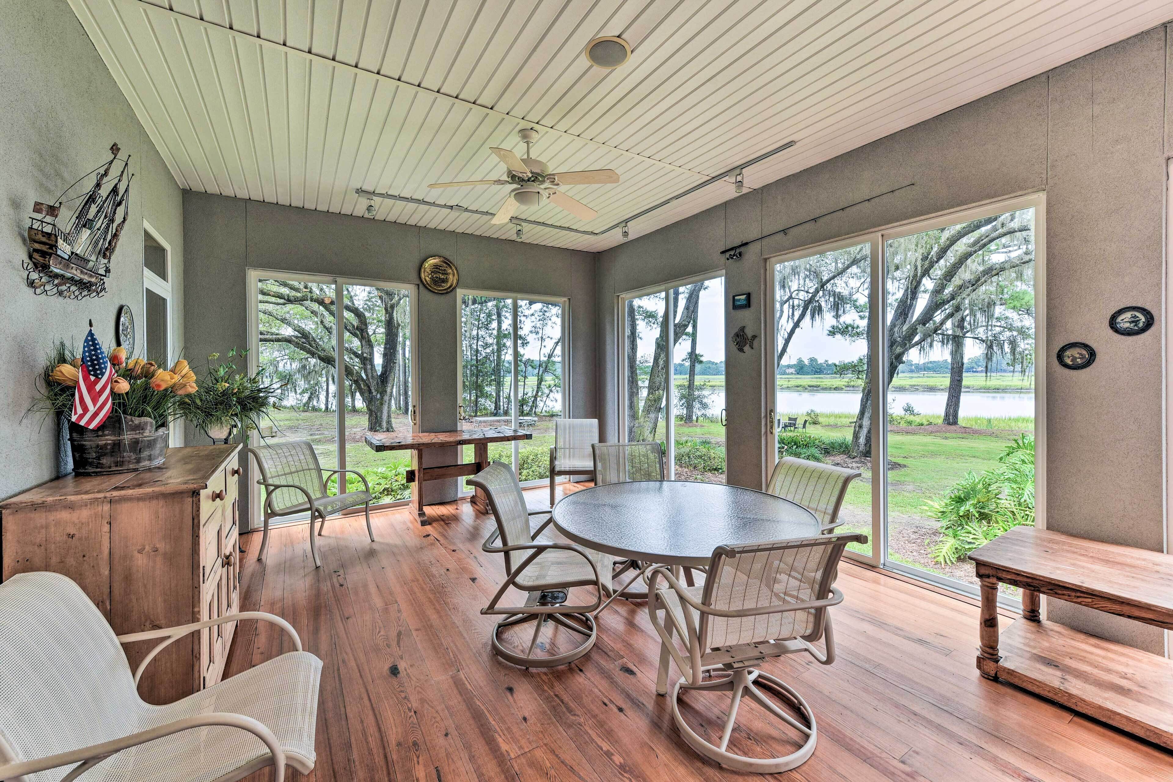 A spacious room with a large round table, comfortable chairs, and a ceiling fan. Large windows offer views of a lake and trees.