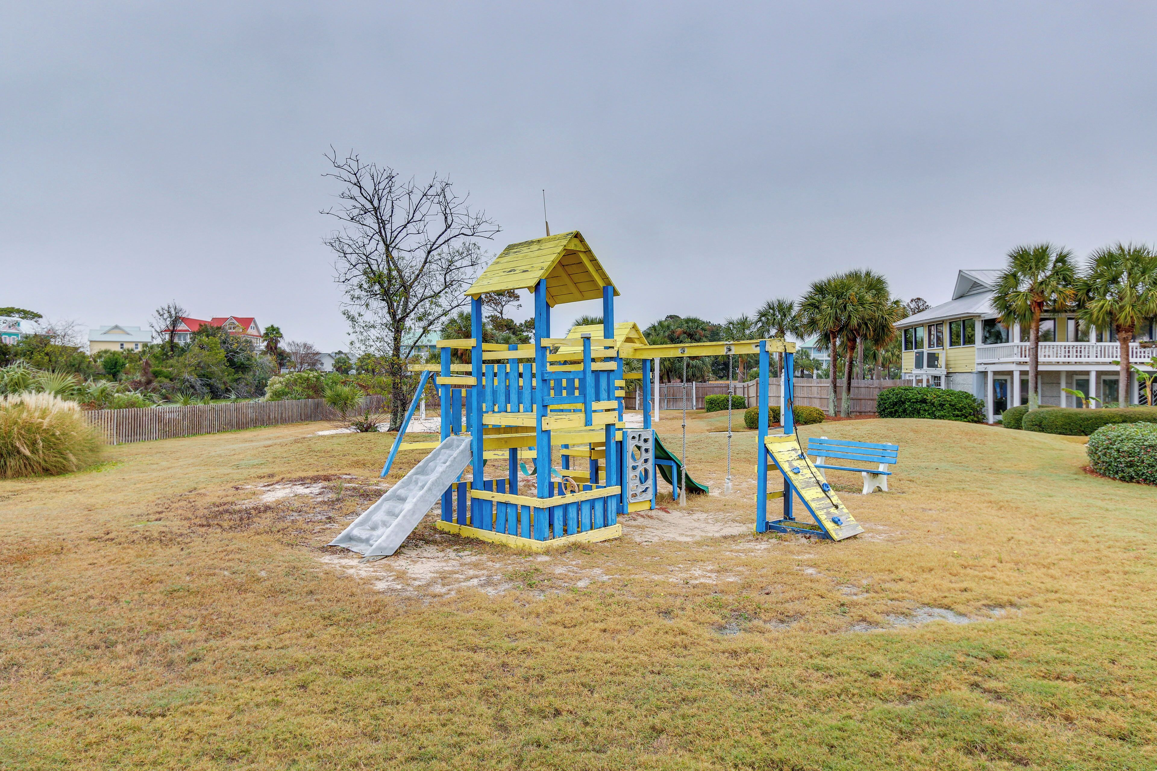 A playground with a blue and yellow play structure, a slide, and a swing set in a grassy area with houses in the background.