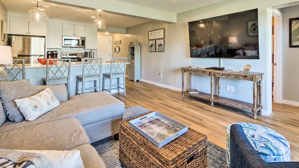 A modern living room with a sofa, ottoman, and a coffee table with a tray. There is a kitchen area with a refrigerator, microwave, and a dining table with chairs. A TV is mounted on the wall, and there is a large window. - St Helena Island Condo w/ Pool & Beach Access! (Harbor Island)