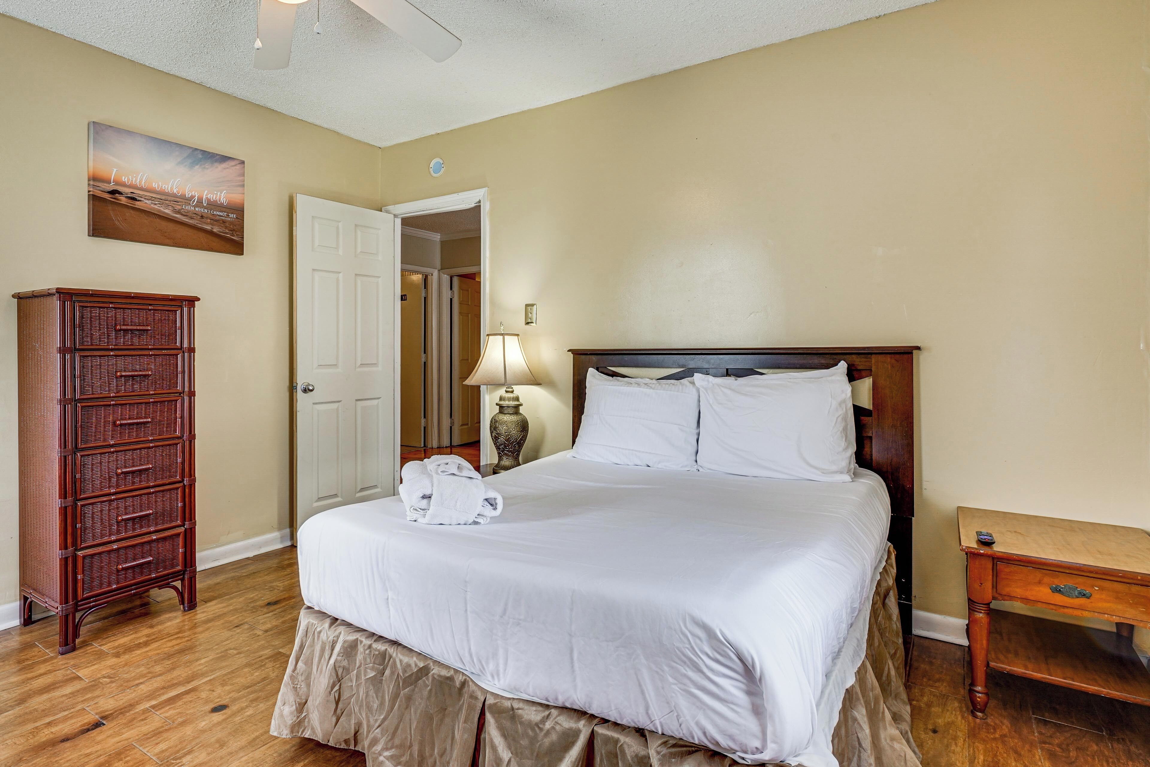 A bedroom with a bed, a wooden dresser, a lamp, and a painting on the wall.