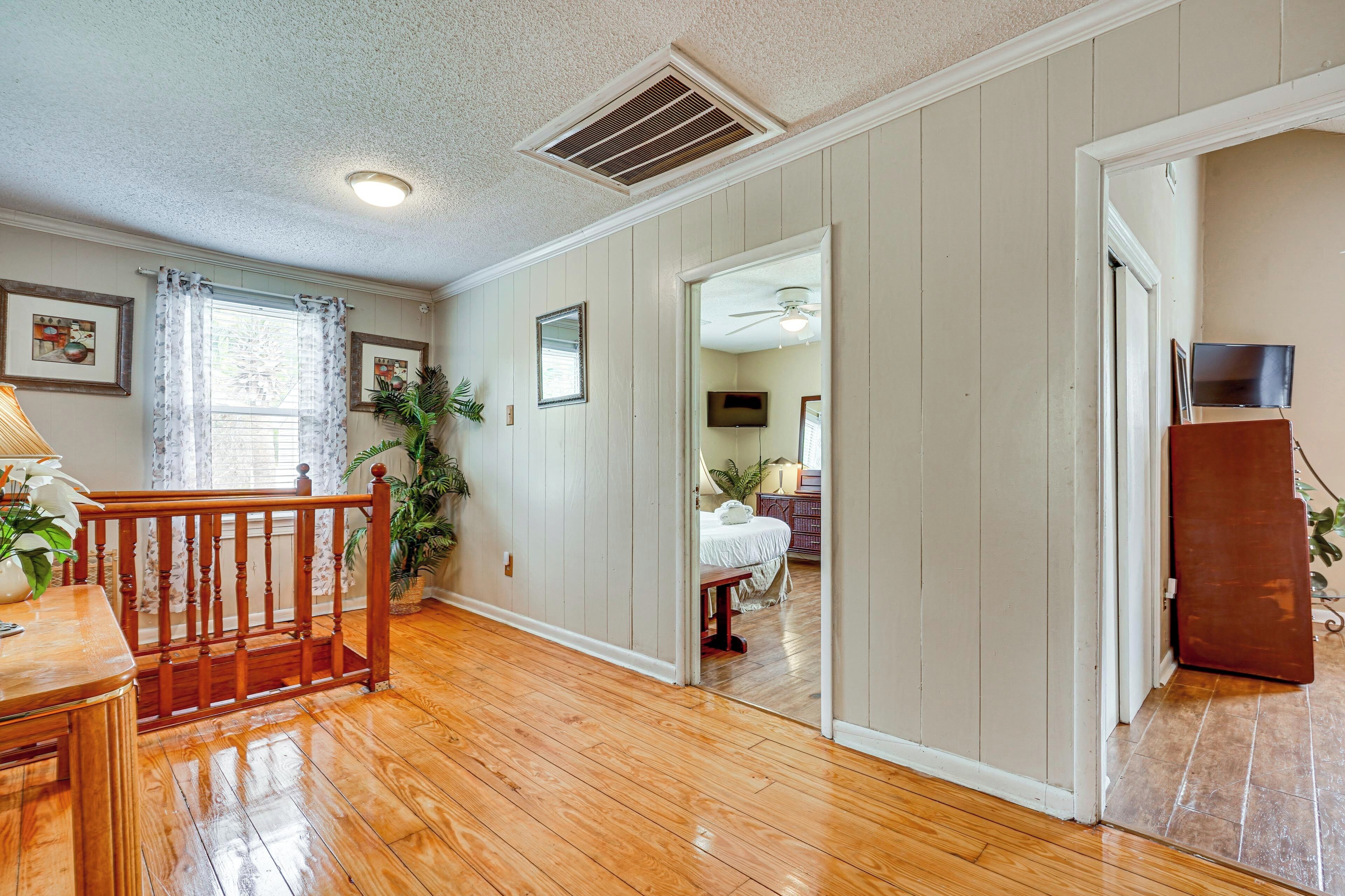 A room with wooden flooring, a staircase, a ceiling vent, and a doorway leading to another room.