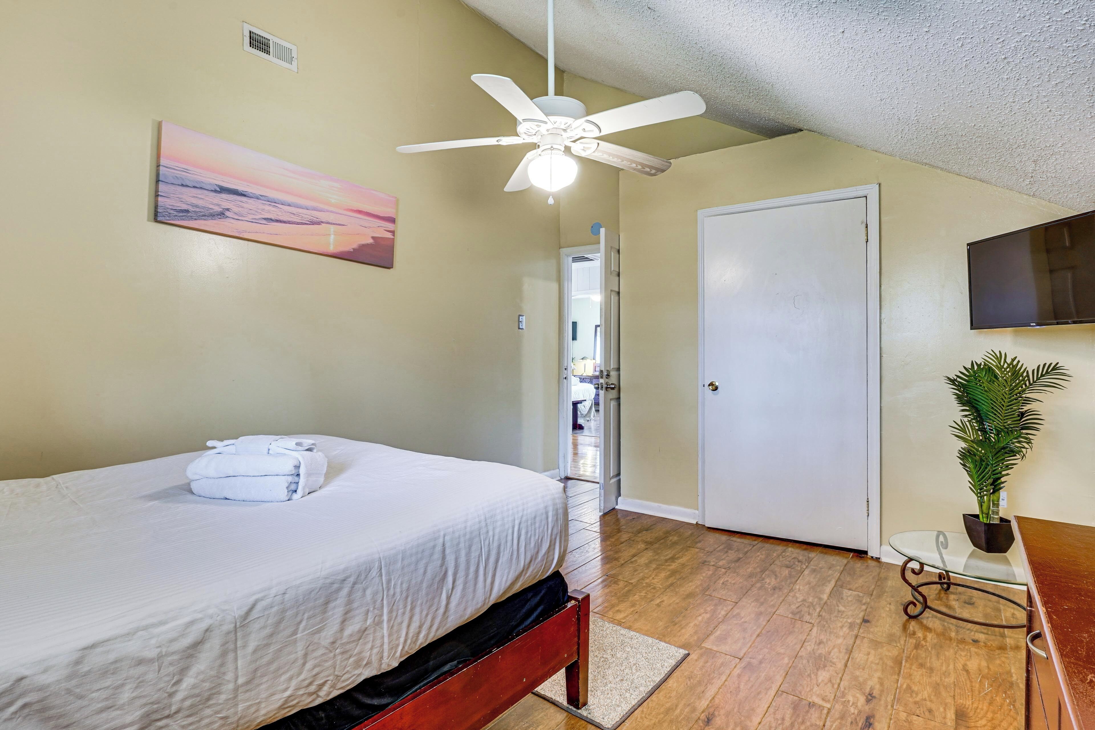 A bedroom with a bed, a ceiling fan, a painting, a TV, and a plant.