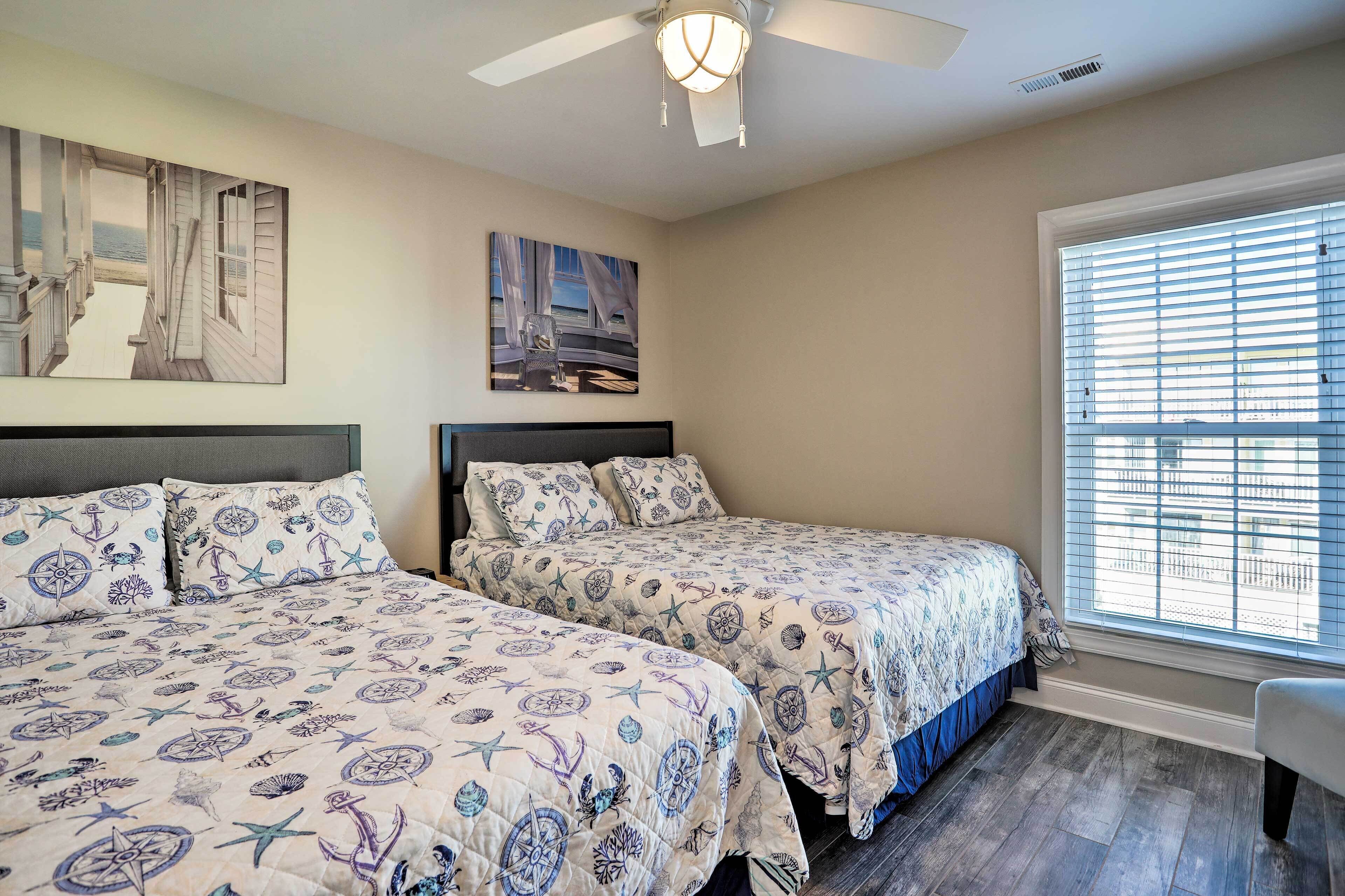 A bedroom with two beds, a ceiling fan, a large window with blinds, and artwork on the walls.