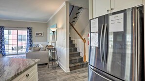 Une cuisine moderne avec un réfrigérateur en acier inoxydable, un escalier avec des rampes en bois et une vue sur l’extérieur par une fenêtre ornée de rideaux à motifs.