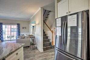 A modern kitchen with a stainless steel refrigerator, a staircase with wooden railings, and a view of the outside through a window with patterned curtains. - Spacious Murrells Inlet Hub w/ Pool, Walk to Shore (Murrells Inlet)