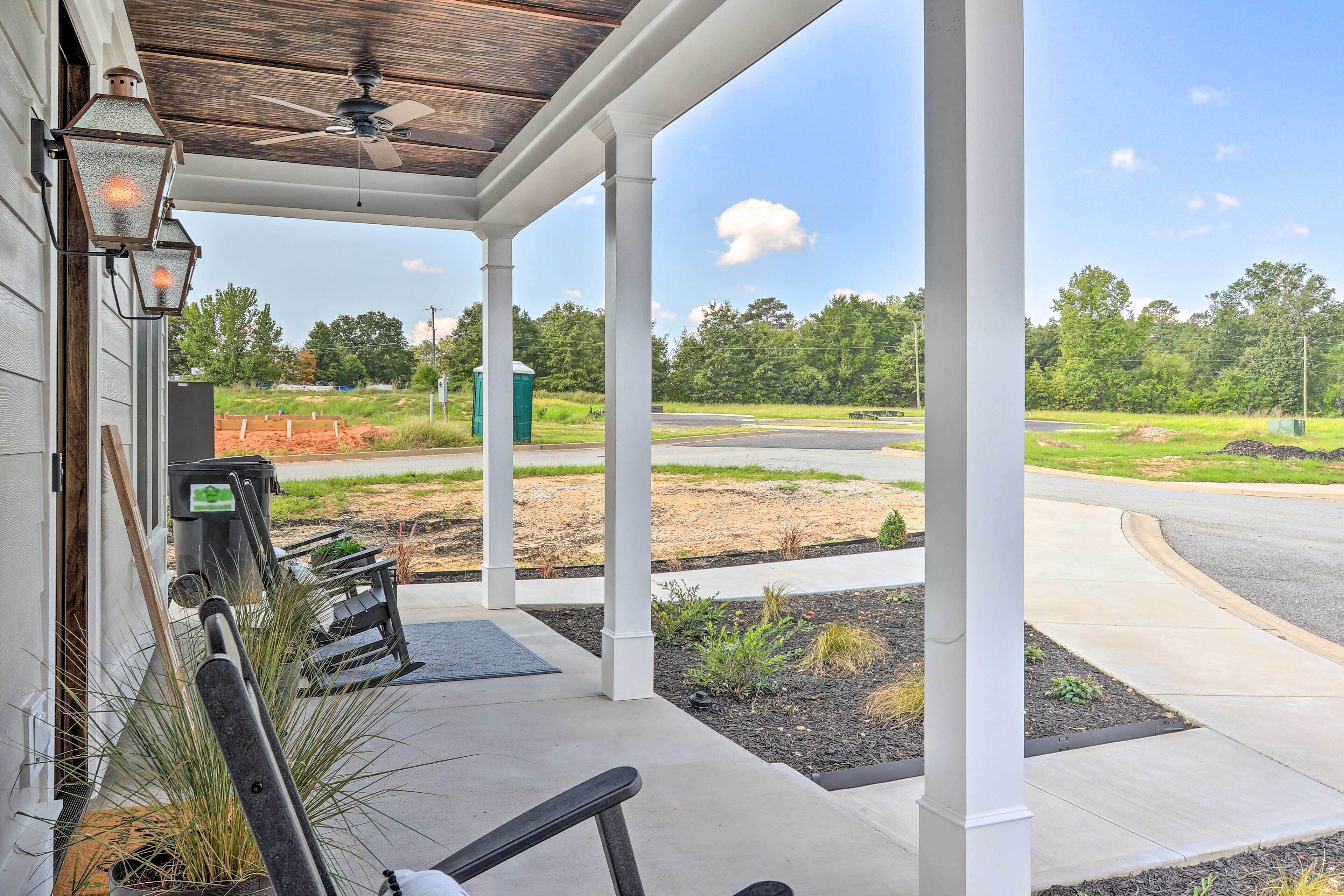 A covered porch with a ceiling fan, outdoor seating, and a view of a landscaped area with a road and trees.