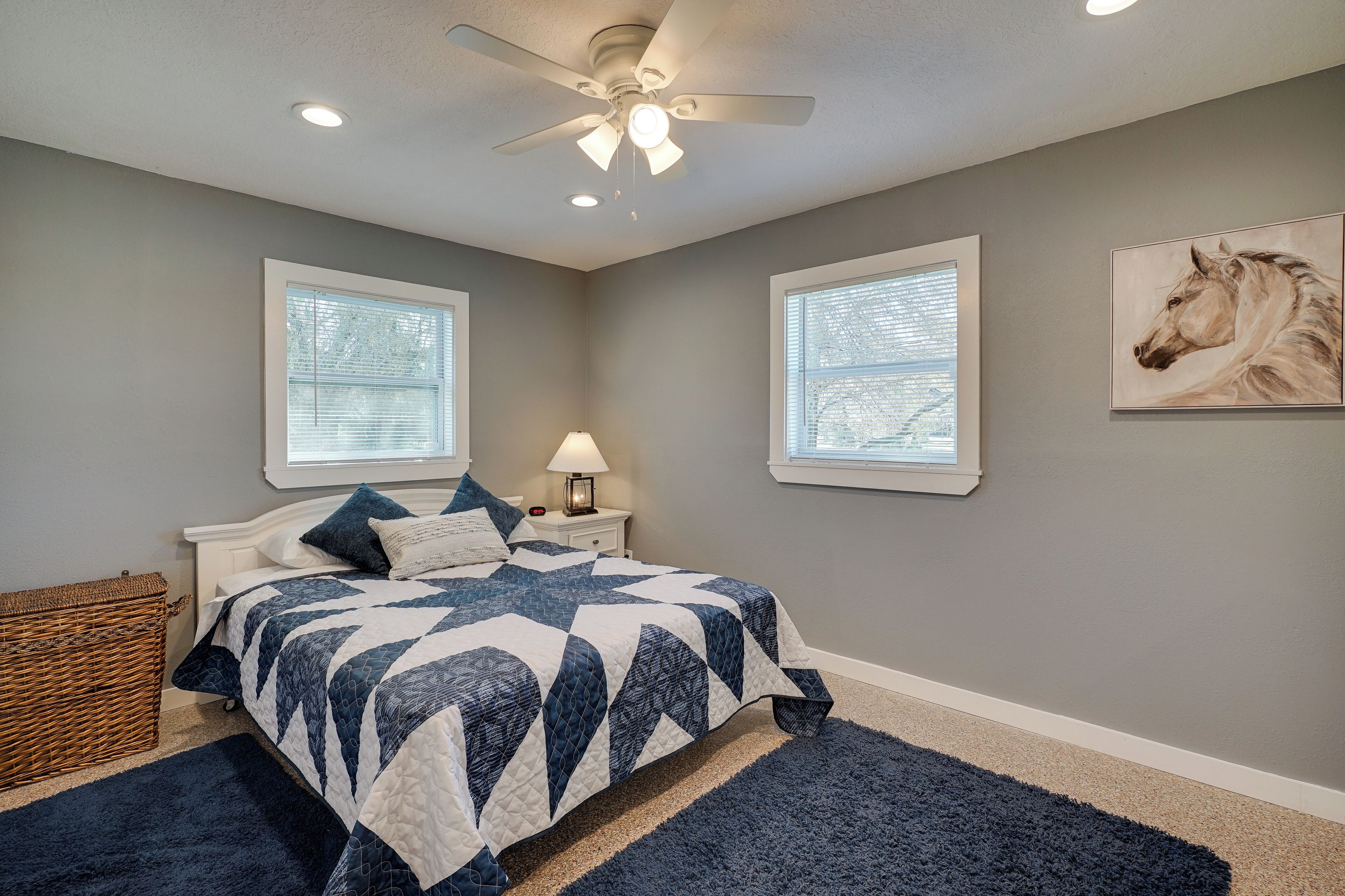 A bedroom with a bed, two windows, a ceiling fan, and a horse painting on the wall.