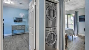 A laundry room with two stacked front-loading washing machines, a small table, and a fan. - Stylish Bayfront Townhome < 10 Mi to Beach! (Mary Esther)