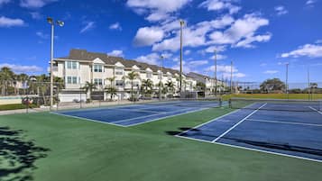 A tennis court with a net, surrounded by a fence, and a building with multiple windows in the background.