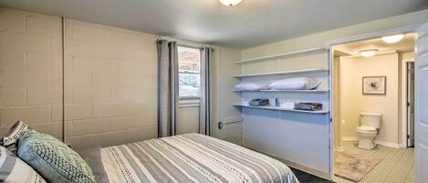 A bedroom with a bed, a window with curtains, a white wall, a door leading to a bathroom, and a shelf with toiletries.