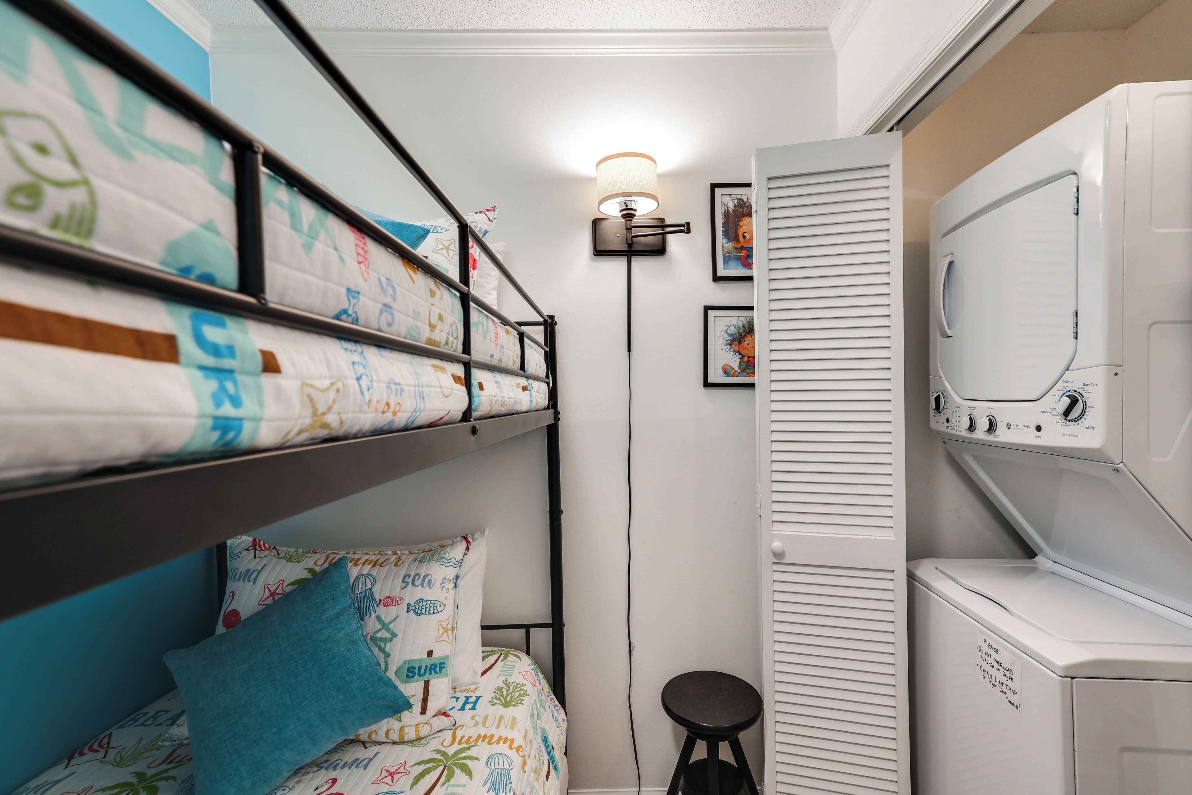A bunk bed with colorful bedding, a standing lamp, a washing machine, and a small stool.