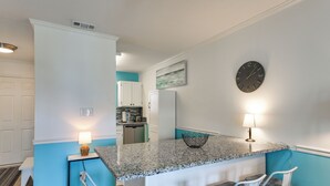 A kitchen with a granite countertop, white bar stools, a clock, and a small island with a bowl. - Across the Street From Ocean: Surfside Beach Condo (Surfside Beach)