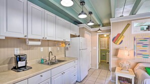 A compact kitchen with white cabinets, a coffee maker, a sink, and a refrigerator. There is a small table with a lamp and a basket.