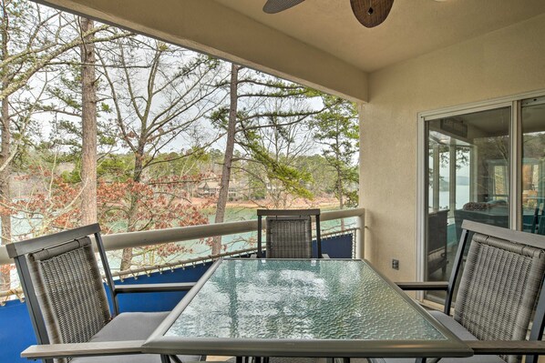 A balcony with a glass table and wicker chairs, overlooking a lake and trees.