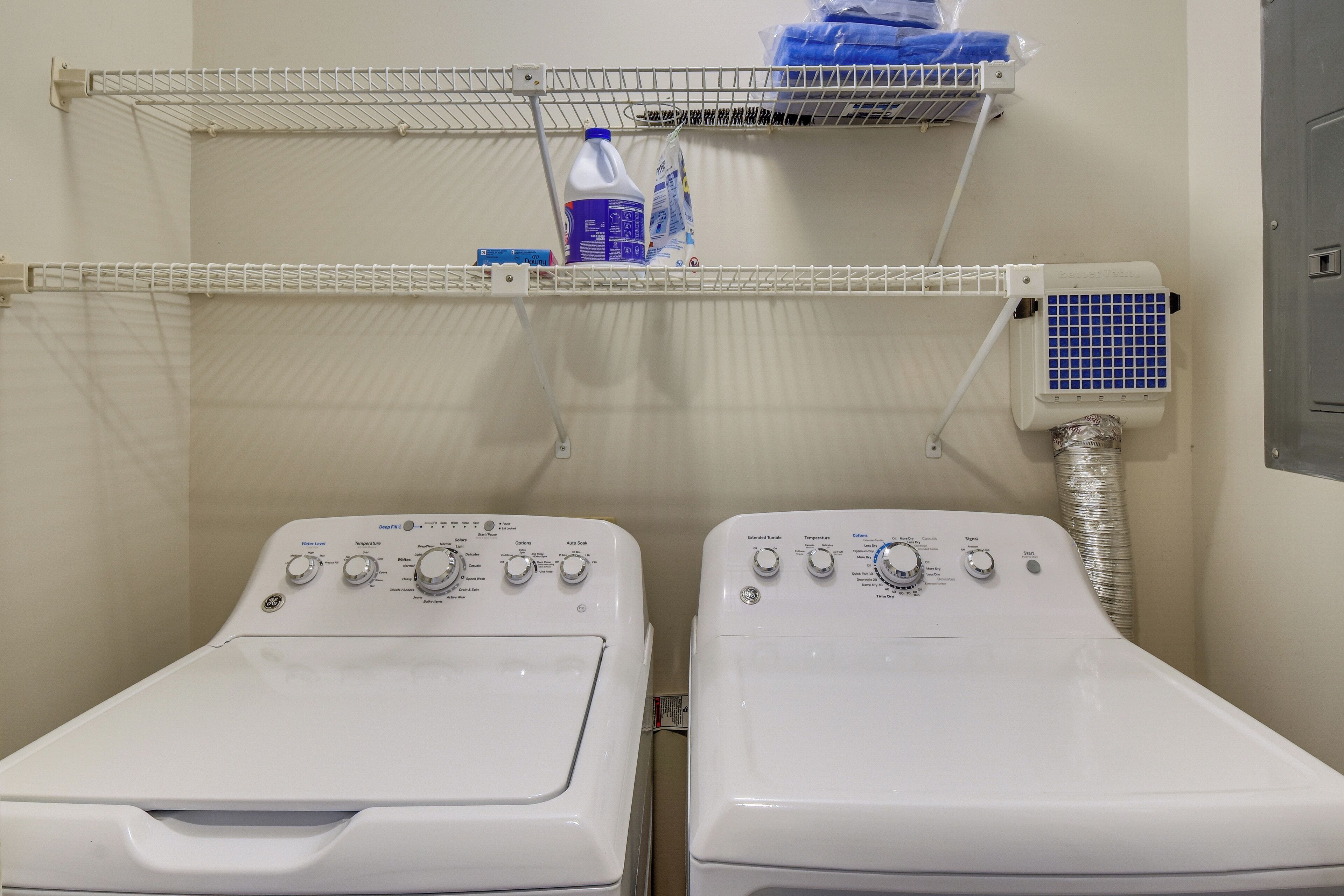 A laundry area with two front-loading washing machines and a shelf above containing cleaning supplies.