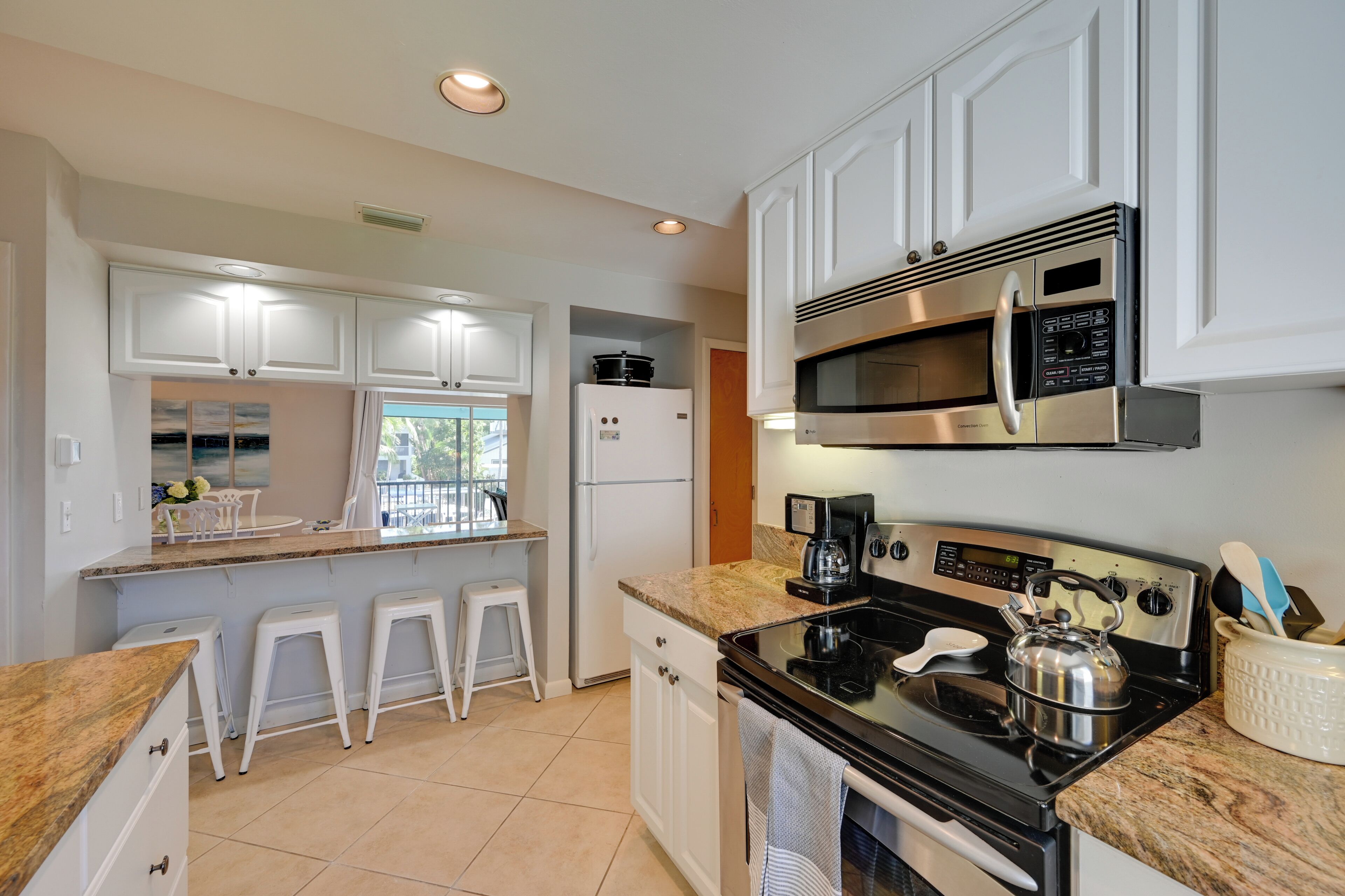 A modern kitchen with white cabinets, a stainless steel microwave, a stove, and a coffee maker. There are two white bar stools and a window with a view of greenery.