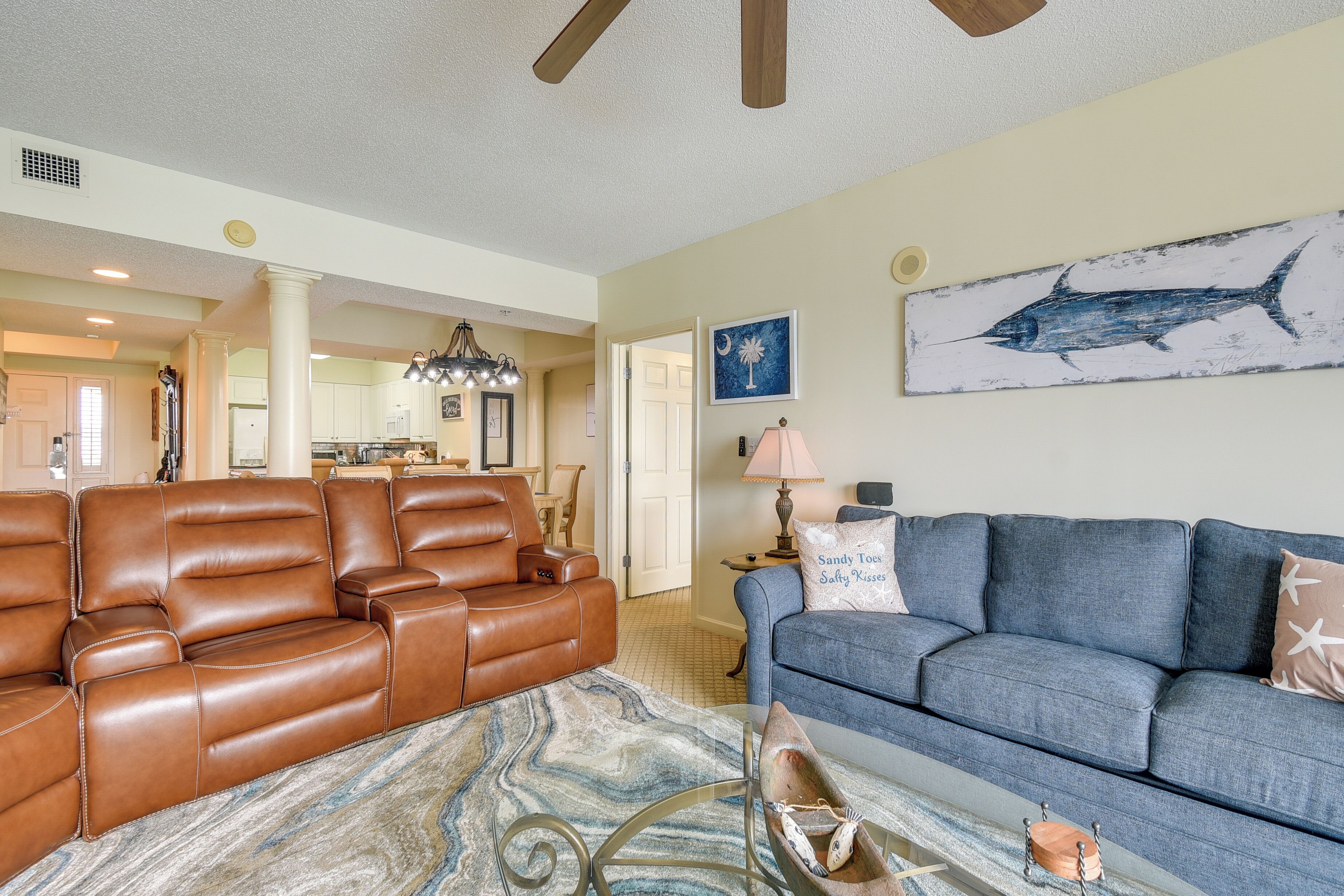 A spacious living room with a brown leather sectional sofa, a blue sofa, and a glass coffee table. There is a large fish painting on the wall and a chandelier above the seating area.