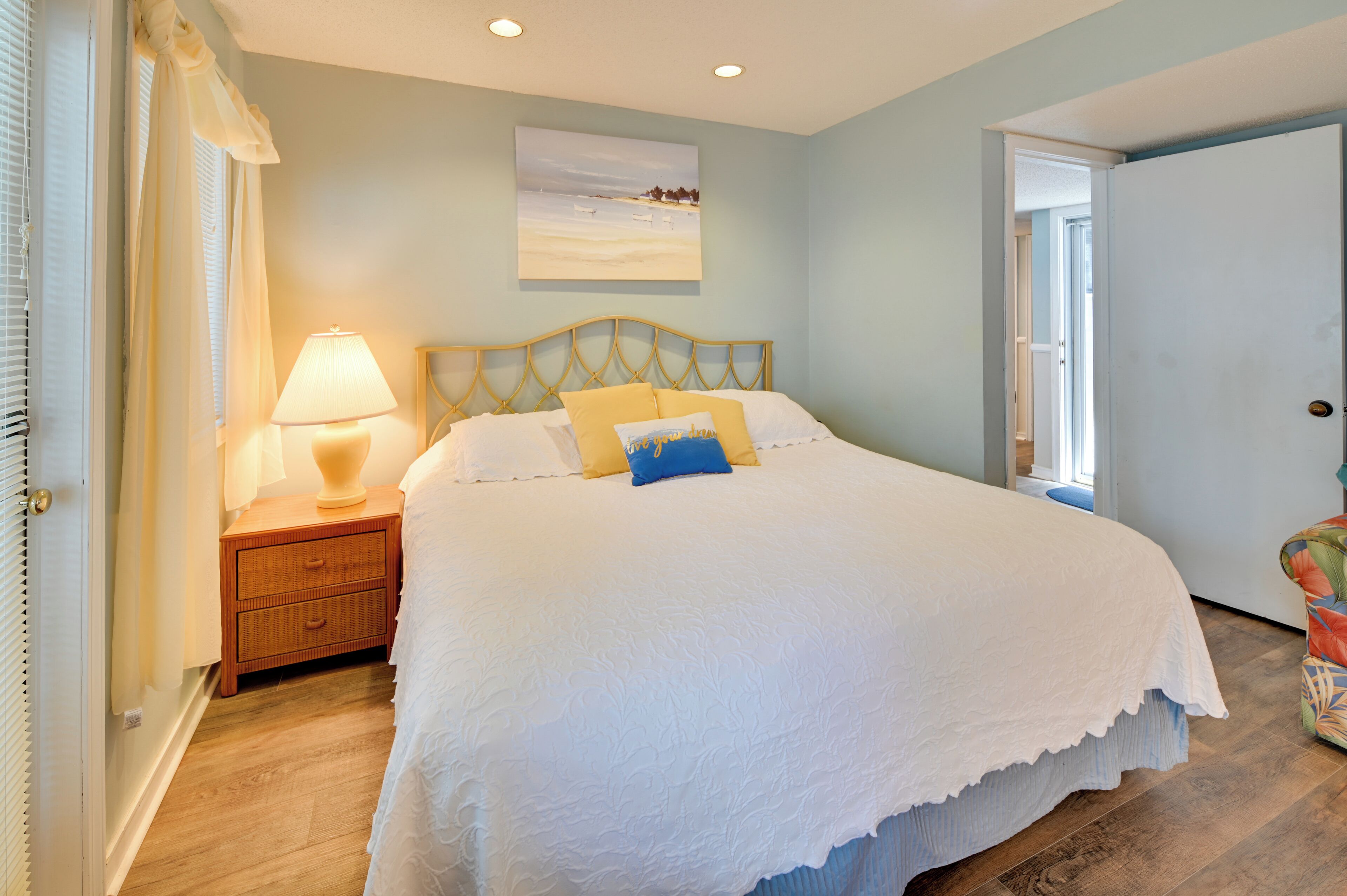 A bedroom with a large bed, a bedside table with a lamp, a window with curtains, and a painting on the wall.