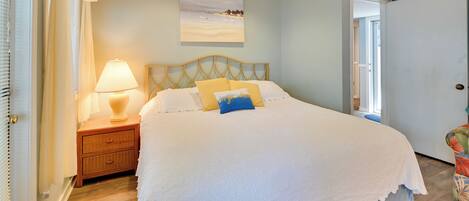 A bedroom with a large bed, a bedside table with a lamp, a window with curtains, and a painting on the wall.