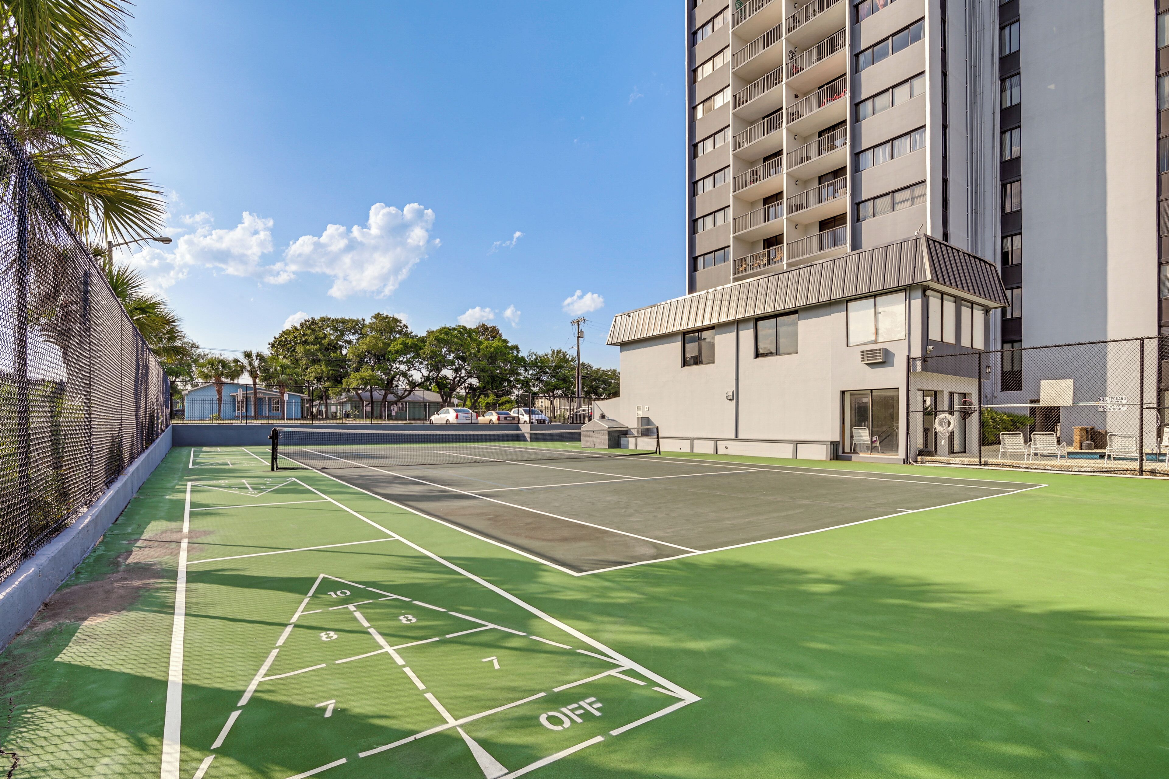 Una cancha de tenis exterior con superficie verde y líneas blancas, rodeada por una valla y edificios.
