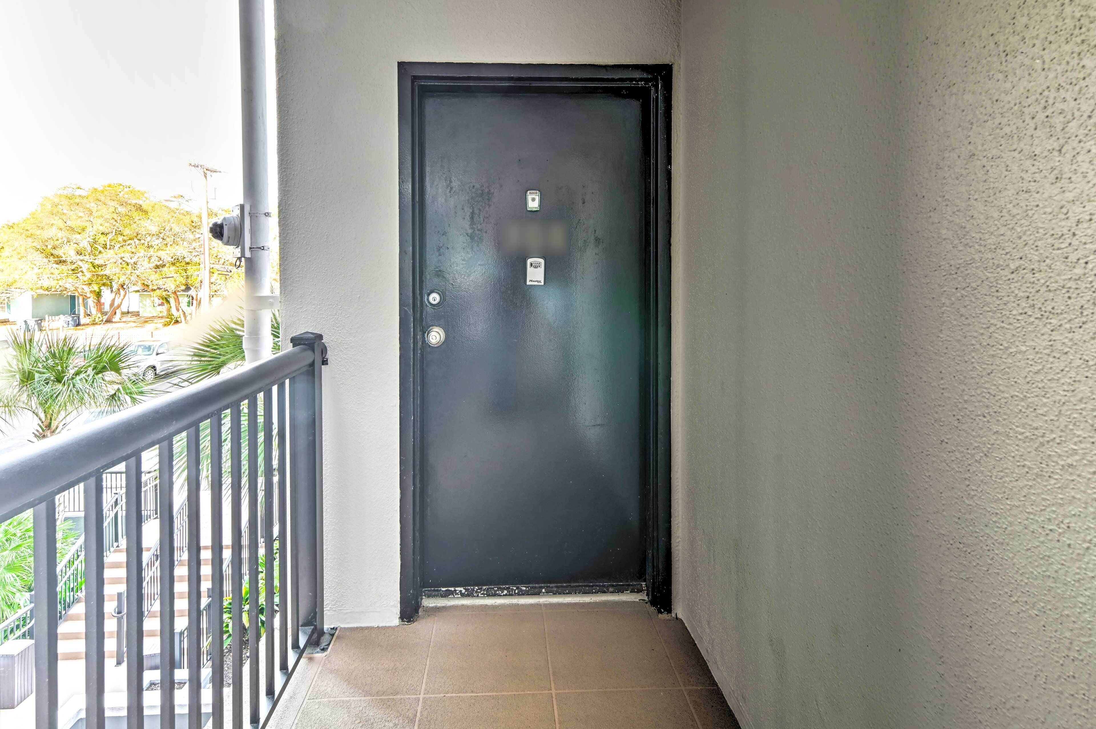 A black door with a security camera and a keypad lock, leading to a balcony with a metal railing and a tiled floor.