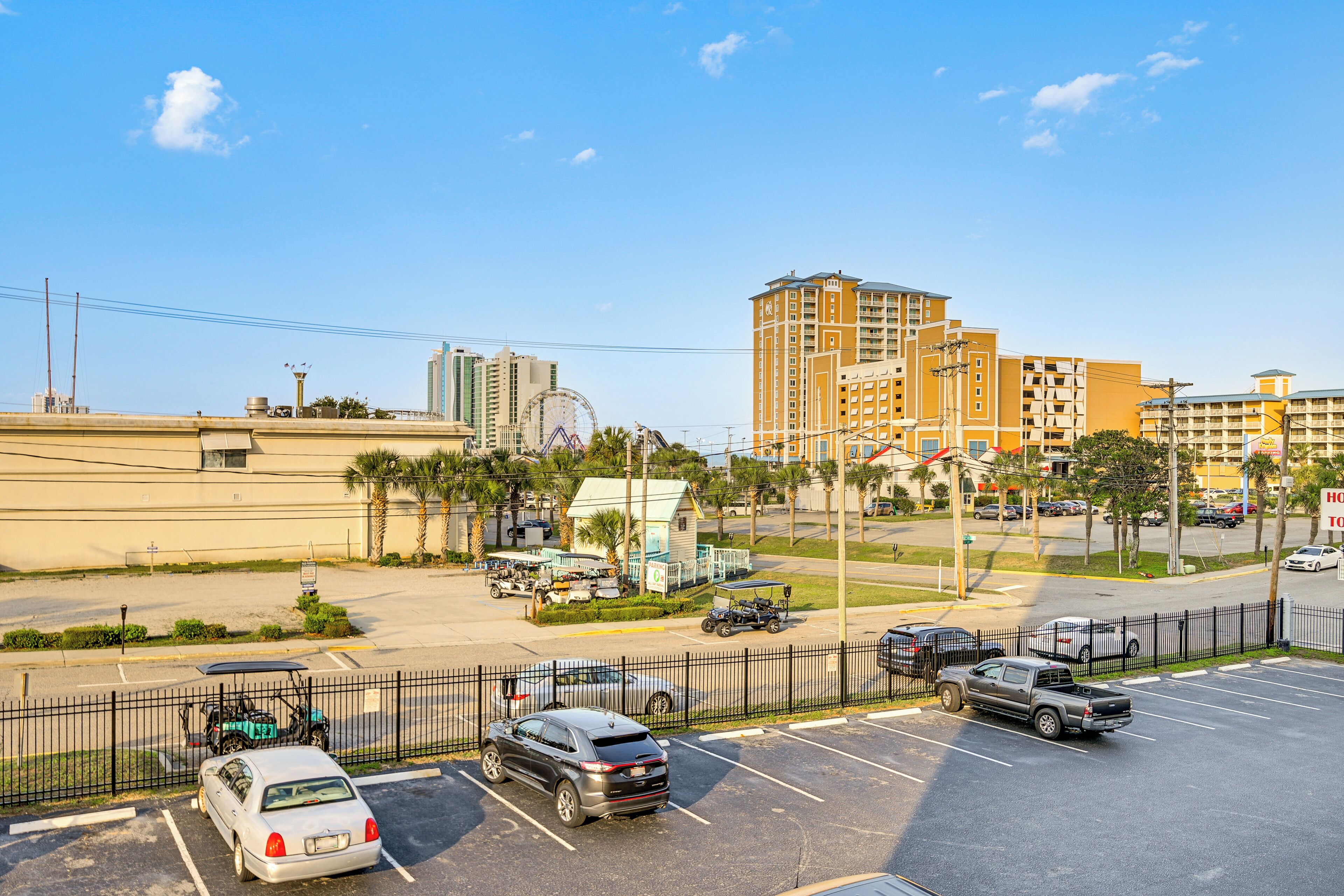 A parking lot with several cars and a golf cart, surrounded by buildings and a Ferris wheel in the background.