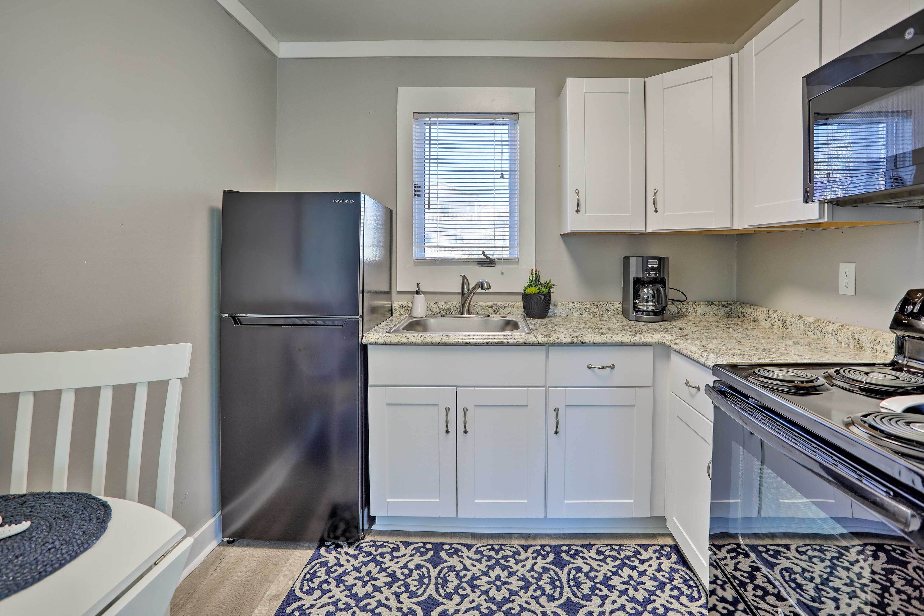 A compact kitchen with white cabinets, a black refrigerator, a window with blinds, a coffee maker, and a patterned rug.