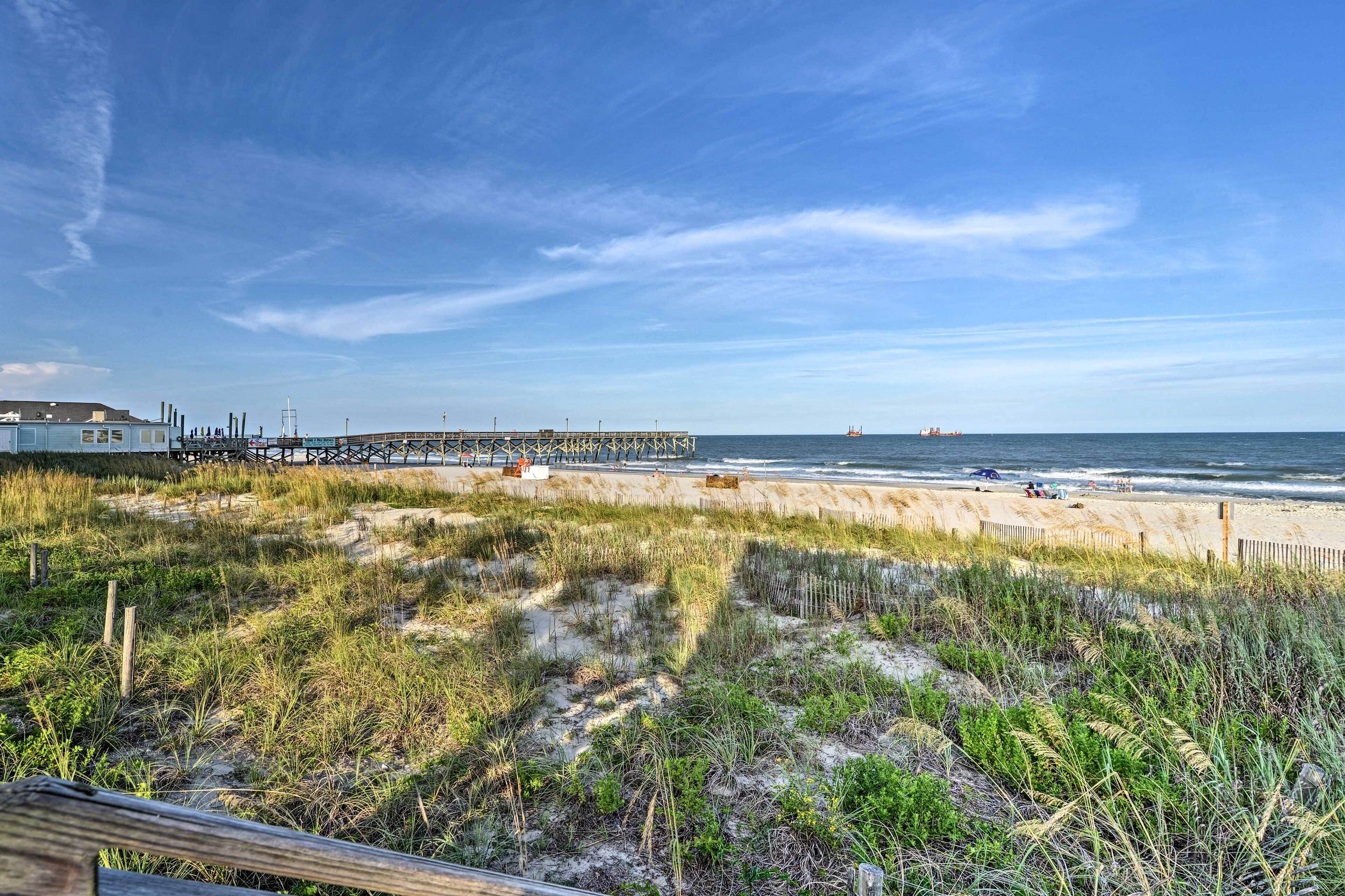 A beach with a wooden pier extending into the water, sandy dunes, and a clear sky.