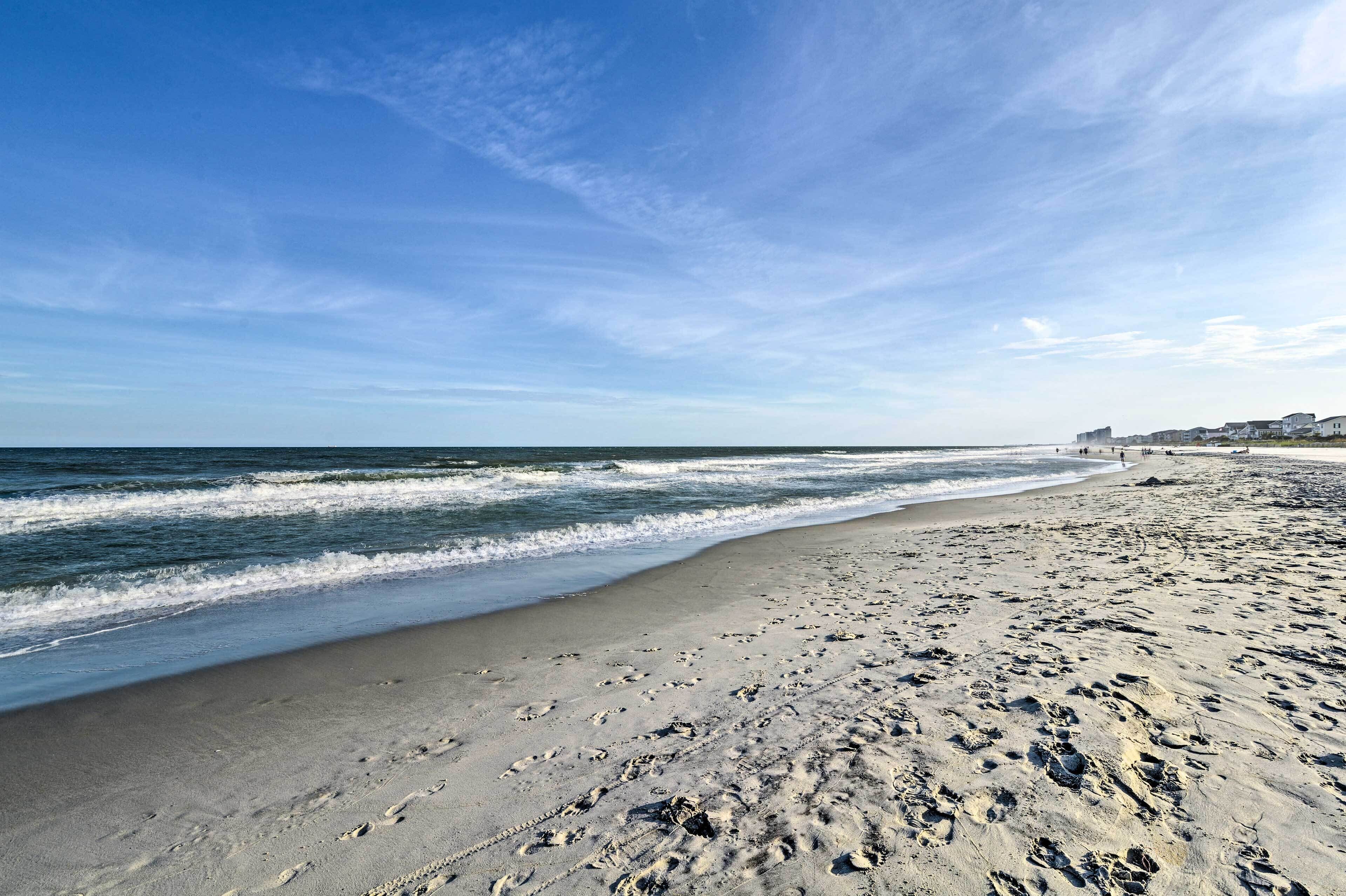 A wide sandy beach with footprints, a clear sky, and a distant shoreline with buildings.